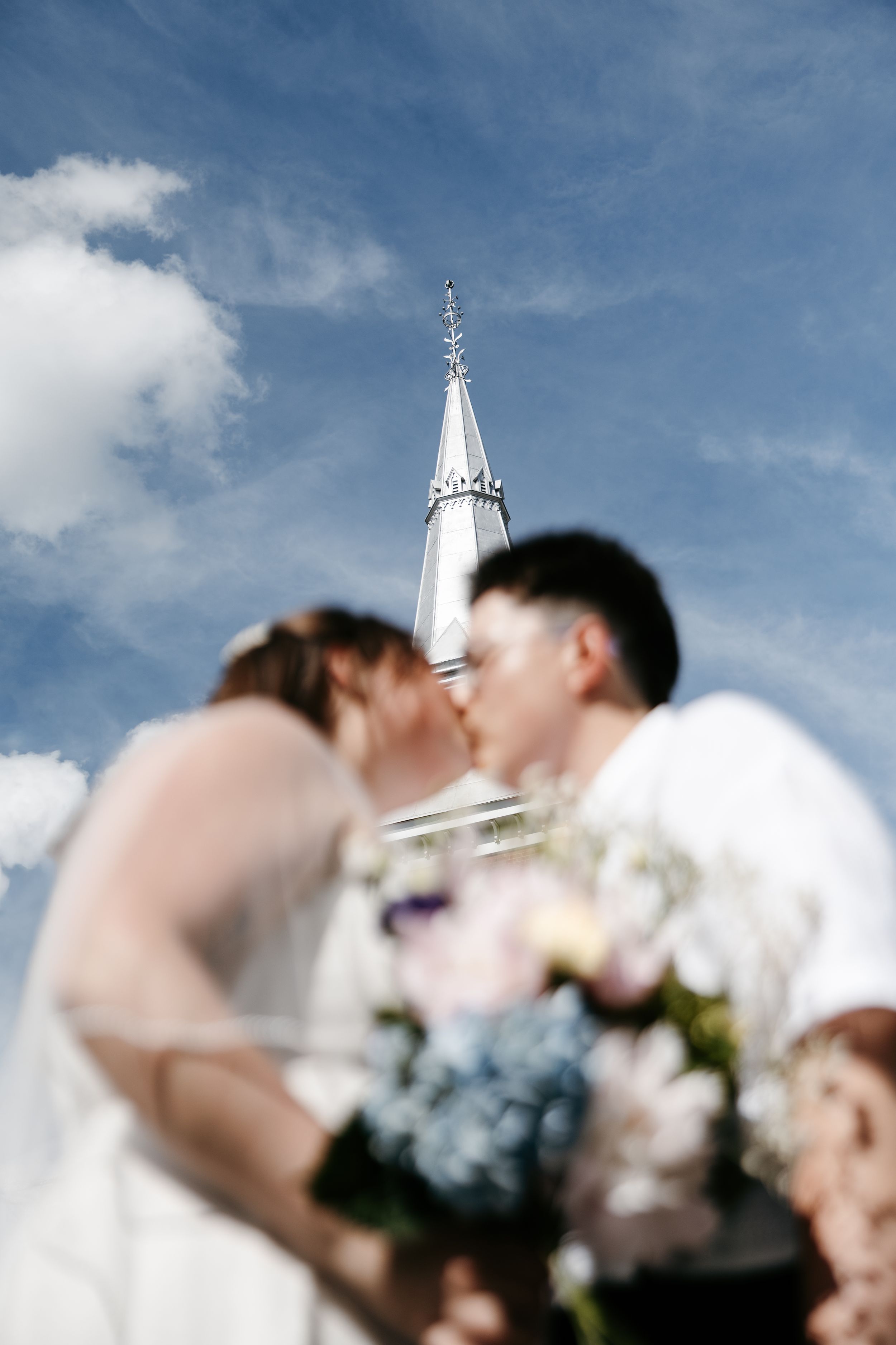 Blurred bride and groom kissing with bouquet in foreground and white church steeple against blue sky.