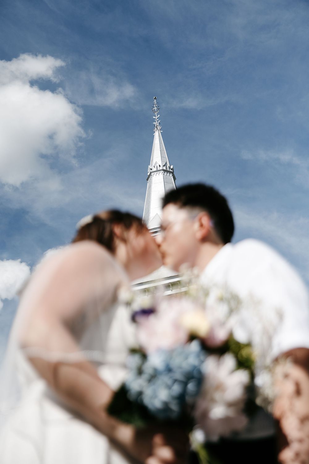 Blurred bride and groom kissing with bouquet in foreground and white church steeple against blue sky.