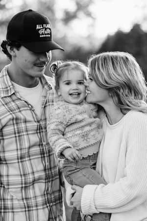 Black and white family portrait of parents holding smiling toddler outdoors, mom kissing child's cheek.