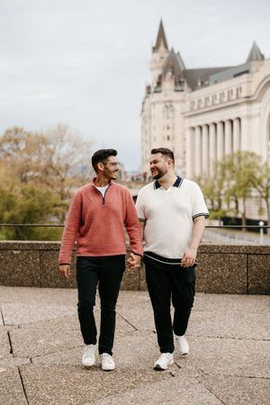 Two men holding hands and smiling at each other while walking outdoors near a historic building.