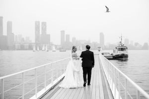 Black and white photo of a bride and groom walking on a pier with a city skyline and boat in the background.