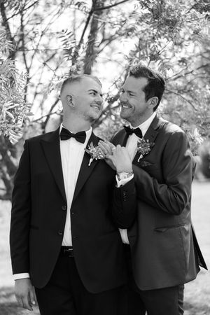 Two grooms in black tuxedos with bow ties and boutonnieres share a joyful moment outdoors in black and white photo.