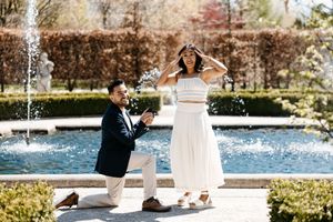 Two people in formal wedding attire hold hands during an outdoor lakeside ceremony under a bright blue sky.