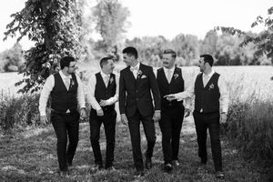 Black and white panoramic view of six groomsmen walking casually together through an outdoor garden setting.