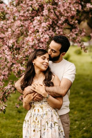 Couple embracing under pink cherry blossom tree, woman in floral dress smiling as man kisses her head in garden.