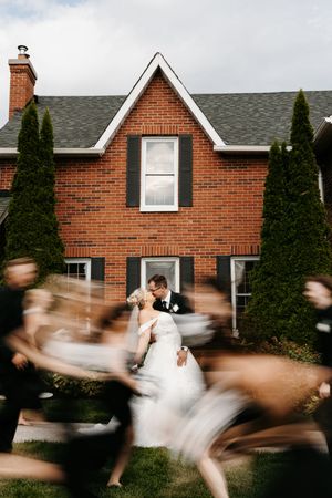 Bride and groom share a kiss outside a brick house while wedding guests rush past in a blur.