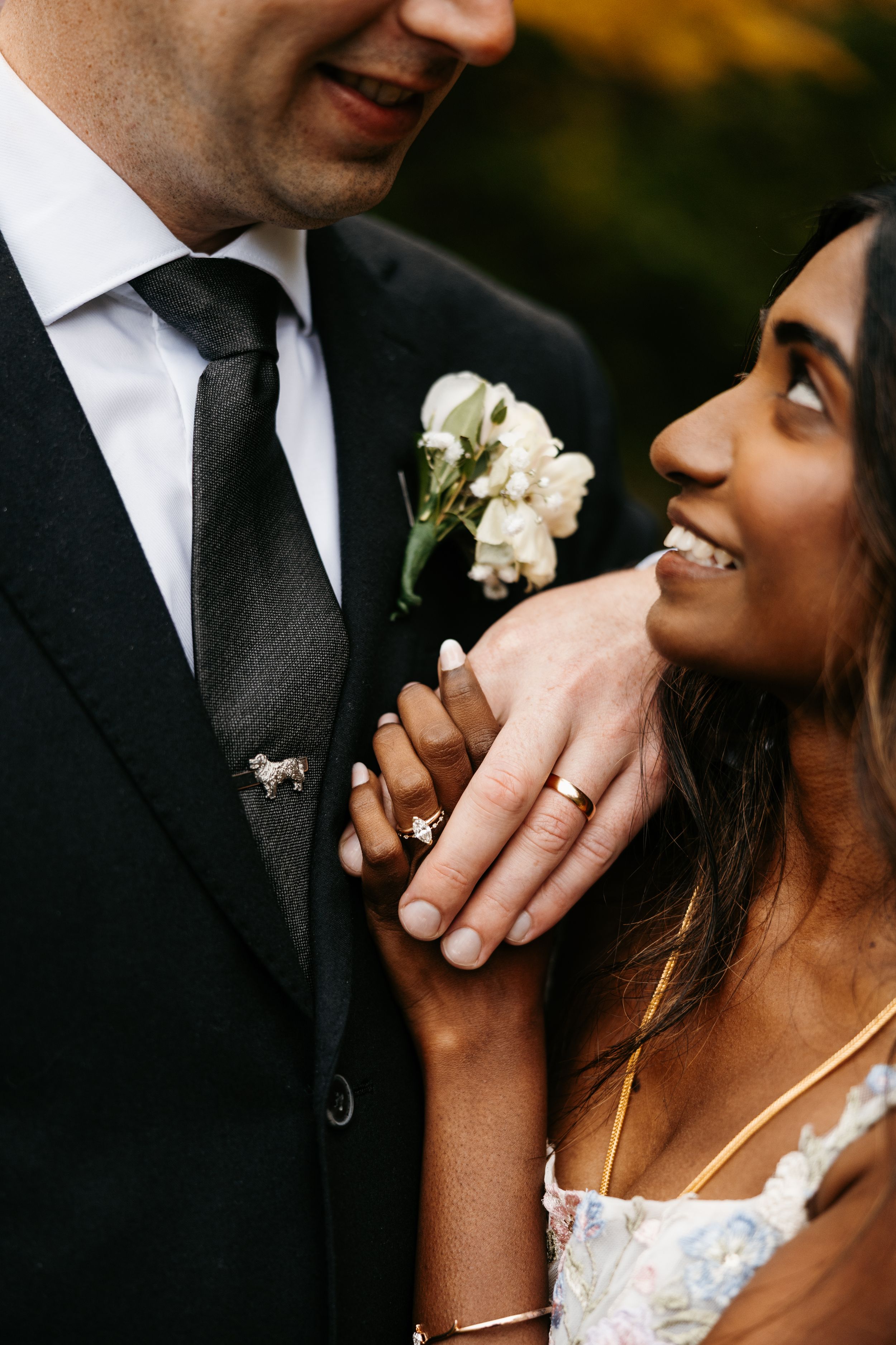Close-up of smiling bride and groom on wedding day, bride wearing floral dress and diamond ring, groom in black suit with boutonniere.