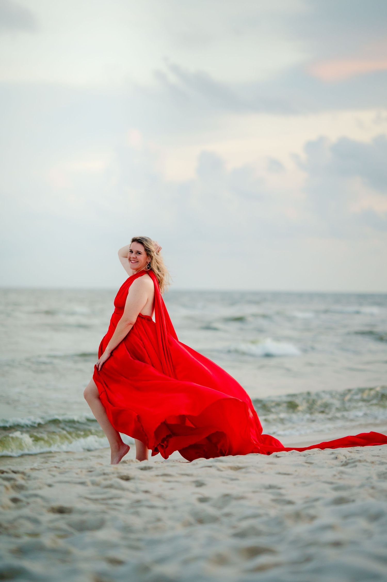 A flowing red dress billows dramatically against the ocean waves and sandy beach at sunset.