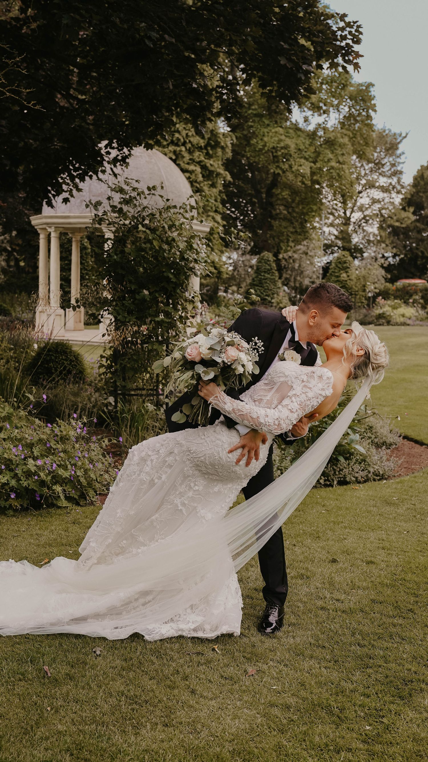 Romantic dip pose on manicured garden lawn with classical gazebo in background.