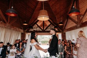 Guests gather under wooden beams and hanging lights at a rustic indoor reception venue during a wedding celebration.