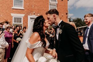 A happy couple sharing a candid moment outside a brick building with guests looking on.