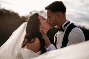 A romantic wedding couple sharing a kiss outdoors during golden hour as the bride's veil flows in the wind.