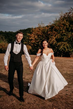 A newlywed couple holding hands outdoors with fall foliage and moody clouds in the background.