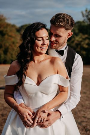 Bride and groom embrace in sunlit outdoor setting as she wears white off-shoulder wedding gown and he wears black tie vest.