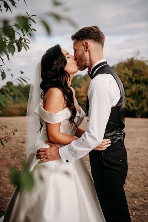 A romantic wedding moment captured outdoors with autumn trees and cloudy skies in the background.