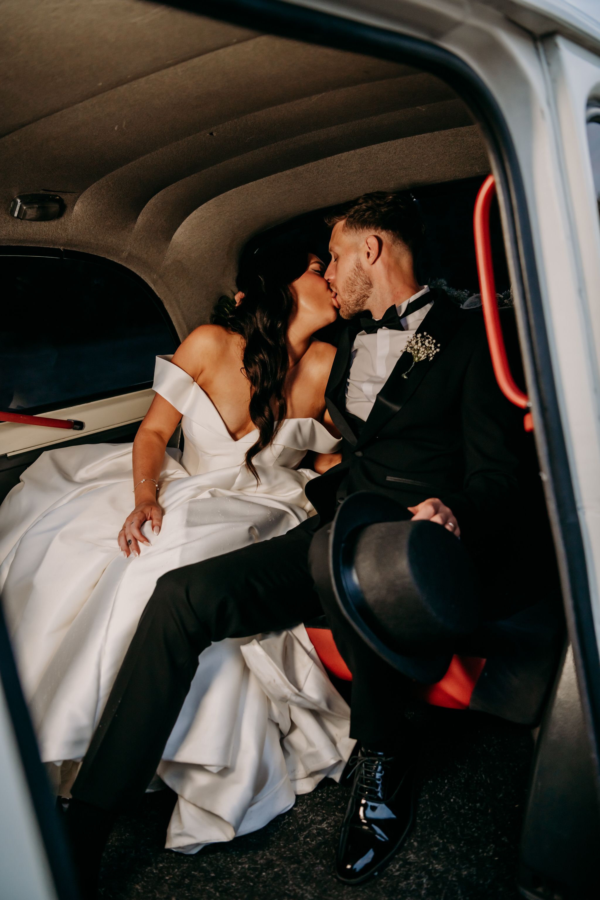 Couple in formal wedding attire sharing an intimate moment in the backseat of a car with dark interior.