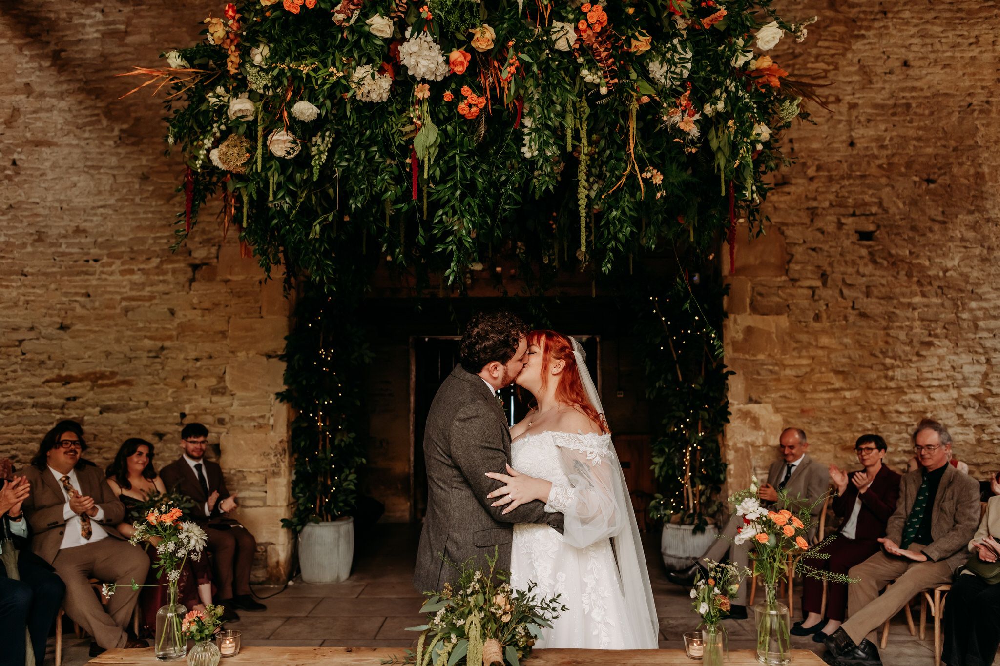 Wedding couple dances surrounded by guests in a rustic brick venue decorated with hanging floral arrangements and greenery.