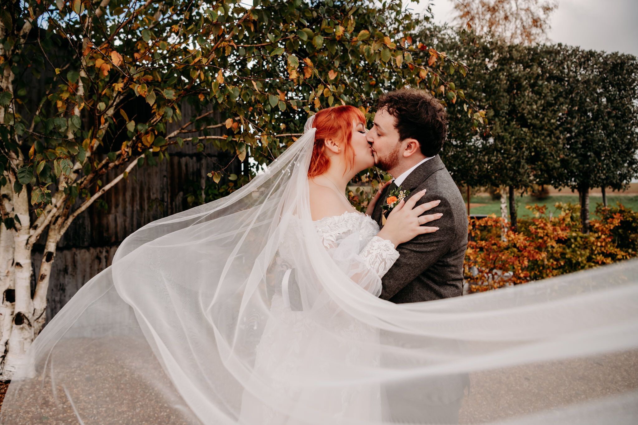 A romantic wedding couple embraces in a garden setting with a flowing white veil catching the autumn breeze.