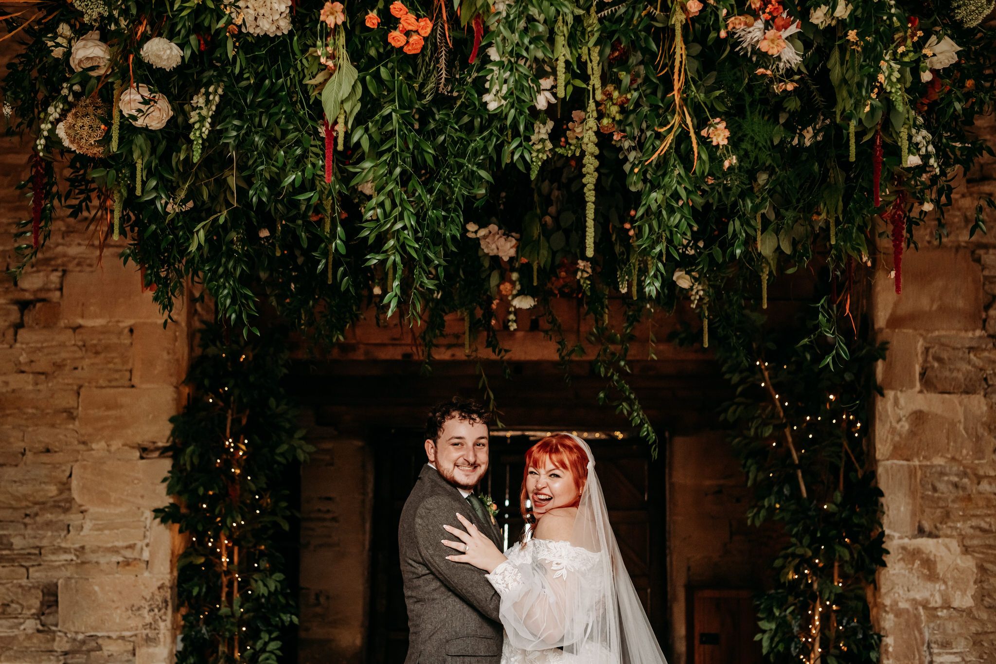 A romantic wedding couple embraces under a rustic stone archway decorated with lush greenery and flowers.