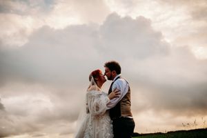 A romantic wedding couple embraces against a dramatic cloudy sunset sky on a hilltop.