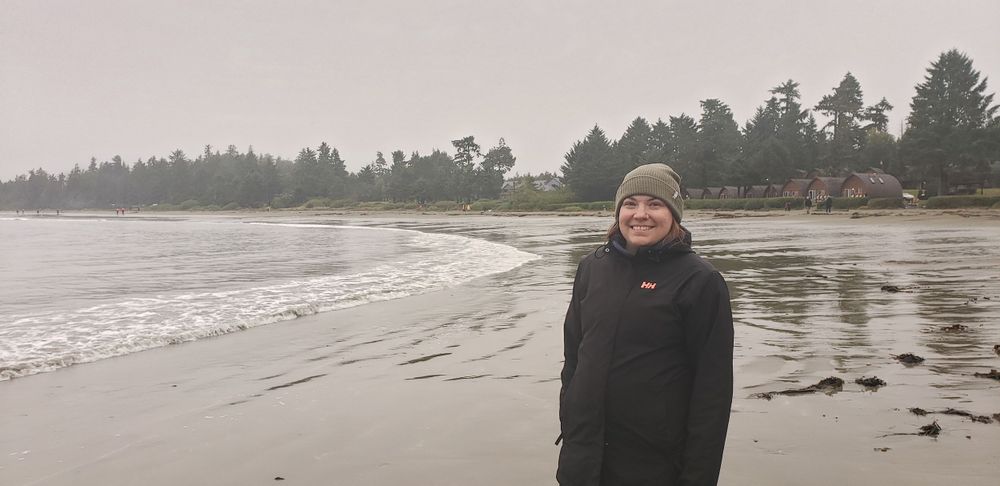 a women stands on a beach, on a foggy day with cabins and trees in the background.
