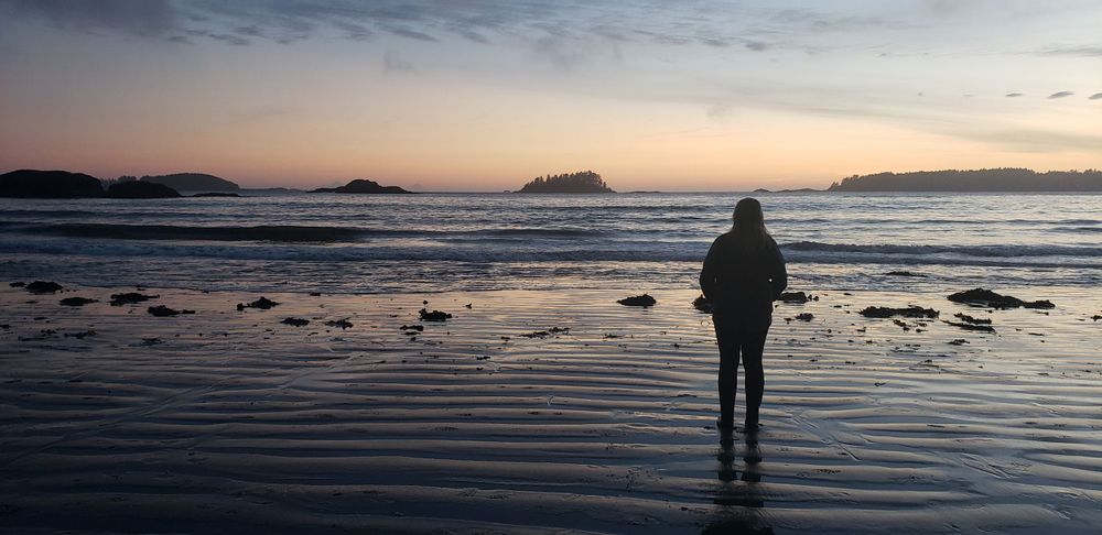 a women stands on a beach looking at the ocean at sunset.