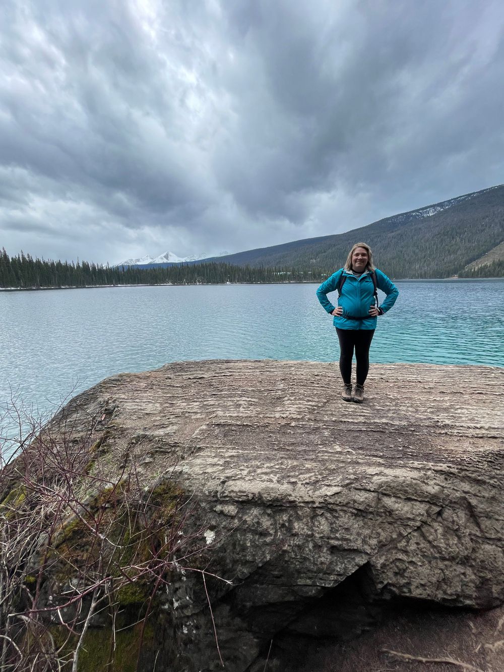 a women stands on a rock by a turquoise lake with mountains in the background.