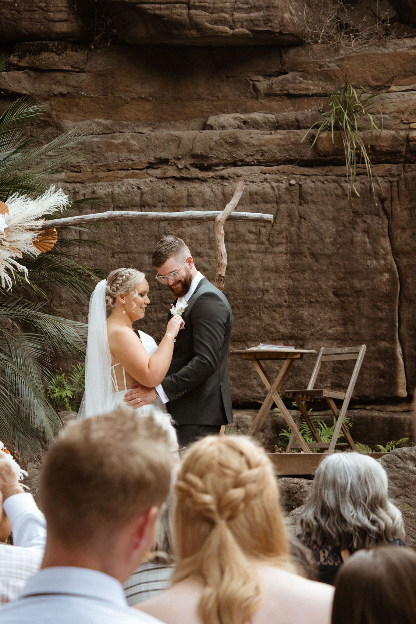 A wedding ceremony takes place against a backdrop of rugged stone cliffs with palm fronds and natural decor.