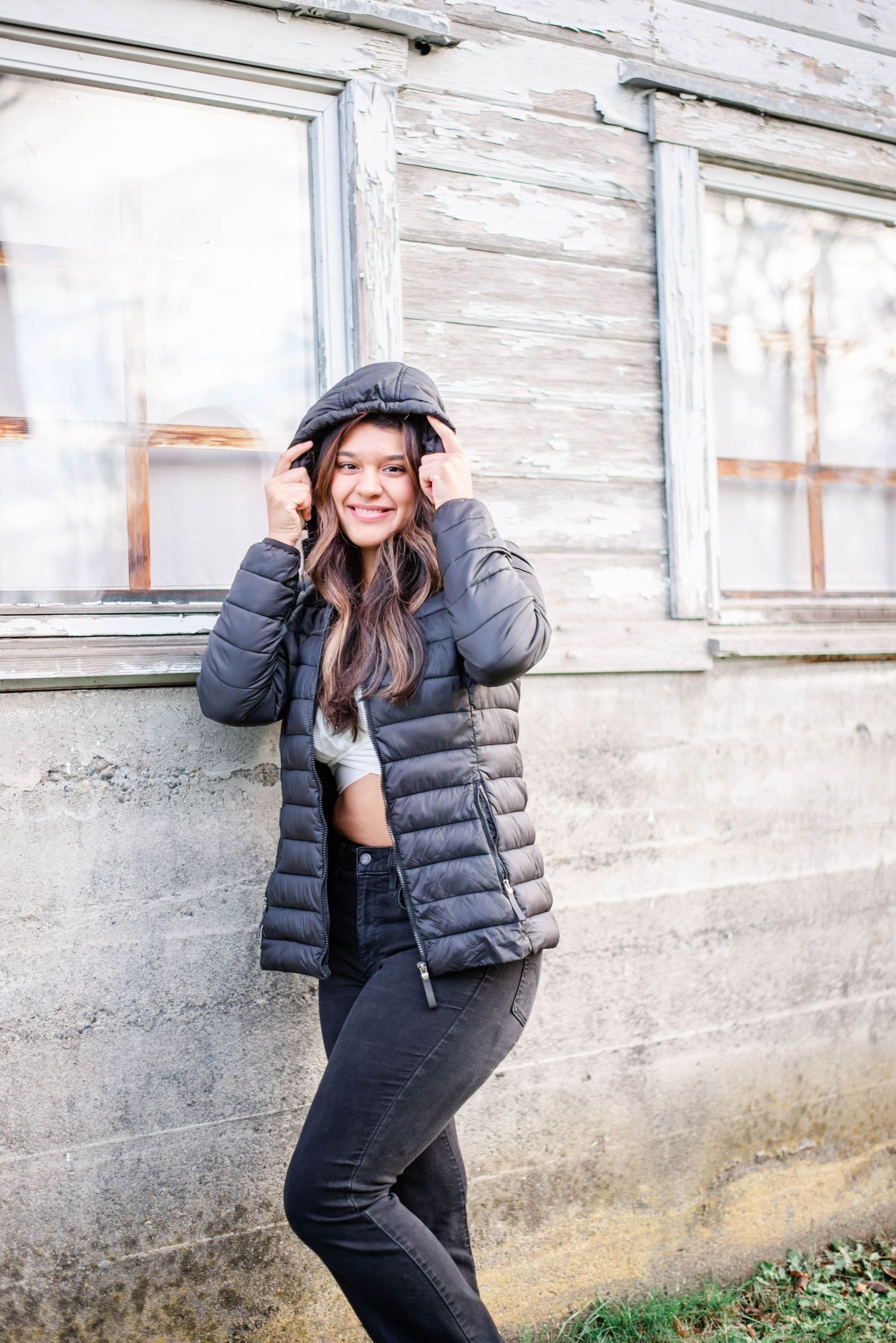 Person in winter attire poses casually against rustic white building exterior.