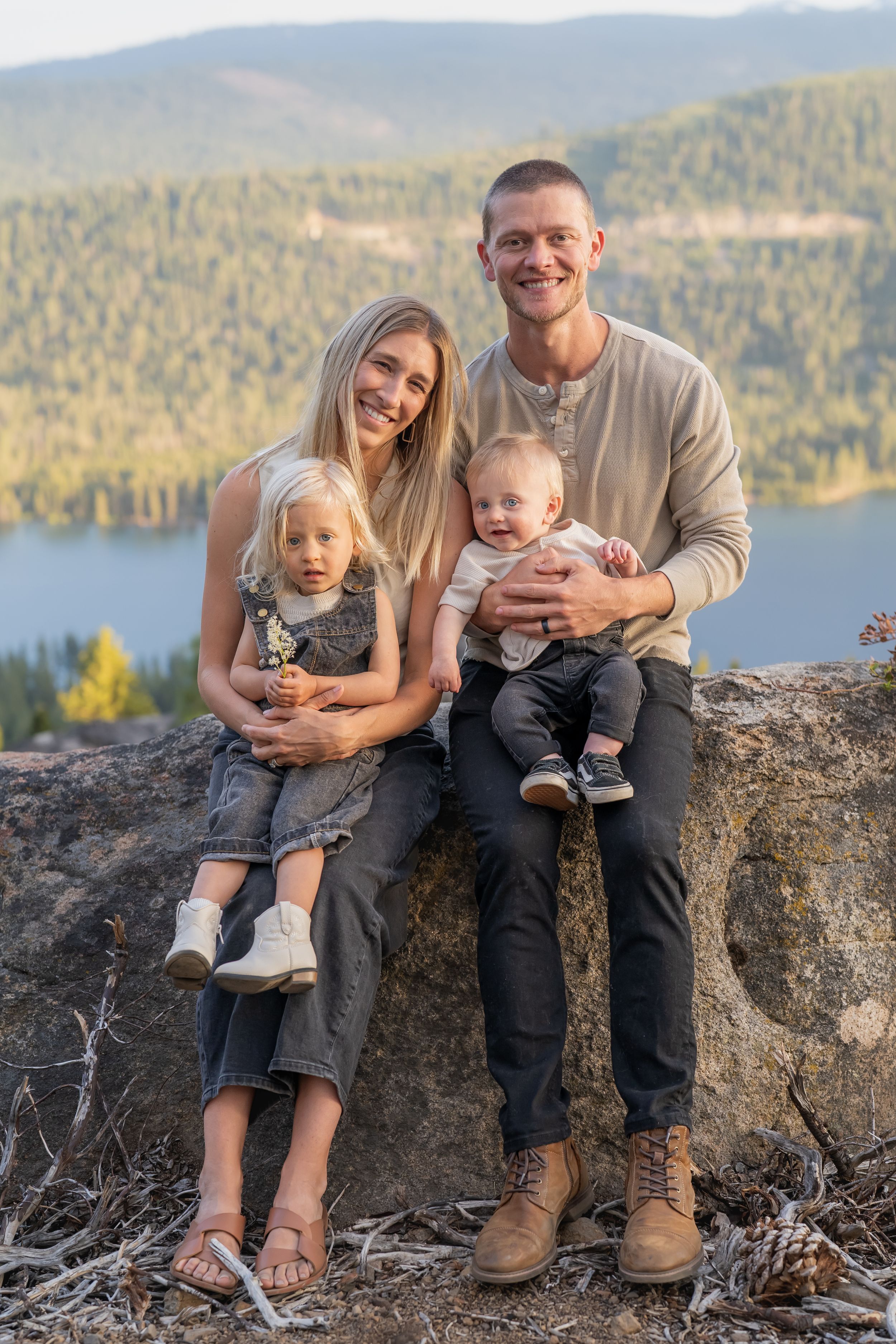 Family sits together on rocky overlook with scenic mountain lake and forest landscape in background during sunset.