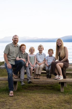 A family sits together on a wooden bench overlooking a serene lake with mountains in the background.