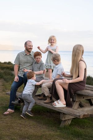 Family sitting together on wooden picnic table at beach during sunset, sharing intimate candid moments.
