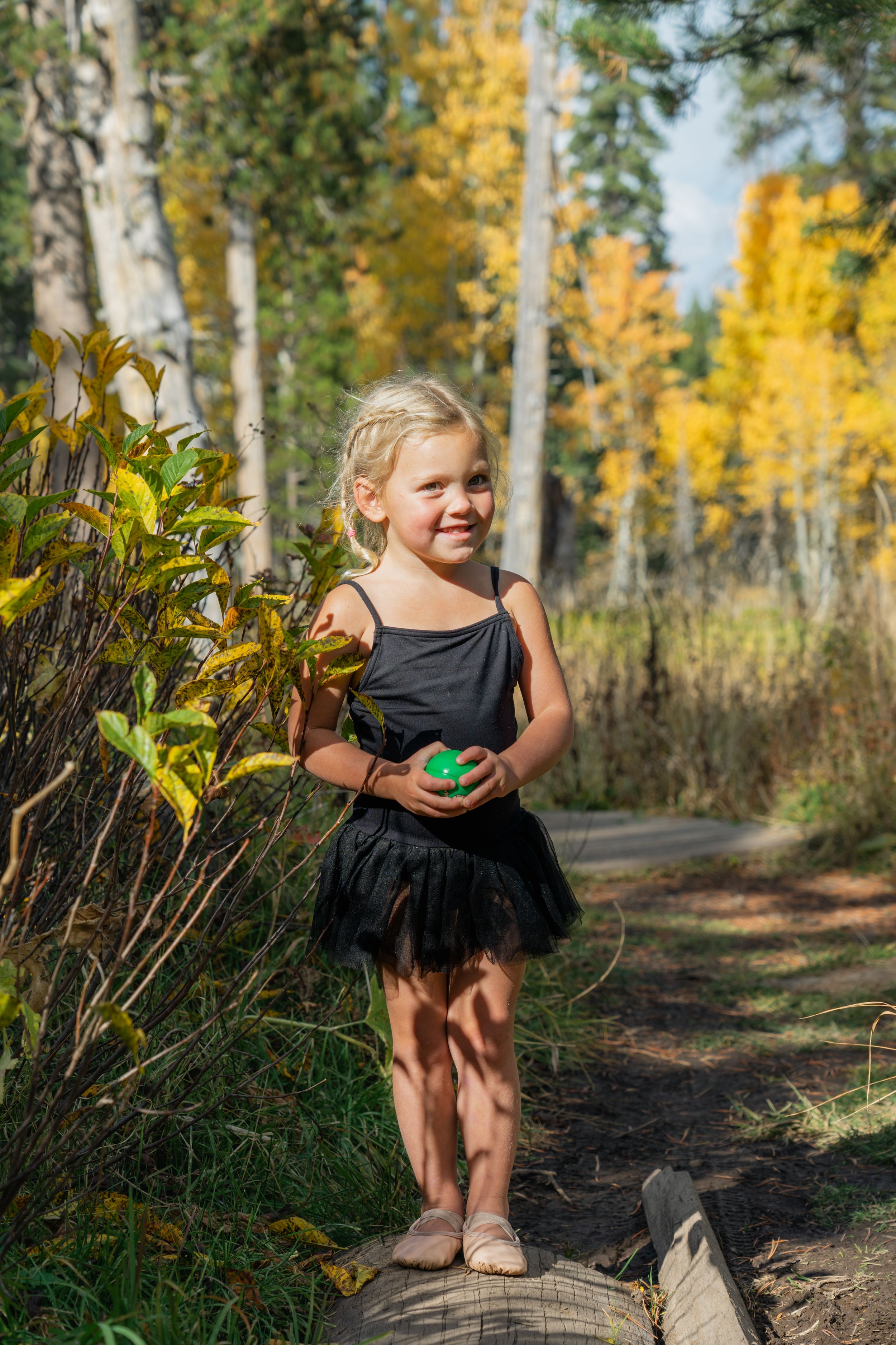 A young dancer in a black tutu dress stands on a log path surrounded by autumn aspens with golden leaves.