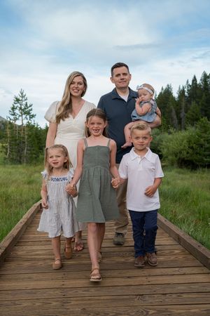 A family poses together on a wooden boardwalk surrounded by green grass and trees in a natural outdoor setting.