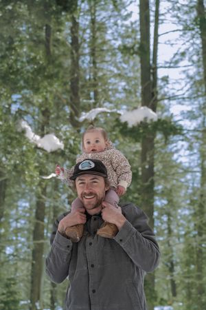 A series of five sequential photos showing a person in a gray jacket and hat playing with a baby in a winter forest setting.
