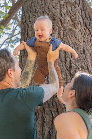A happy baby is lifted up and held against a tree trunk while adults smile and play outdoors.