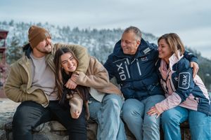 A group of friends laugh together while sitting outdoors in winter against a snowy mountain backdrop.