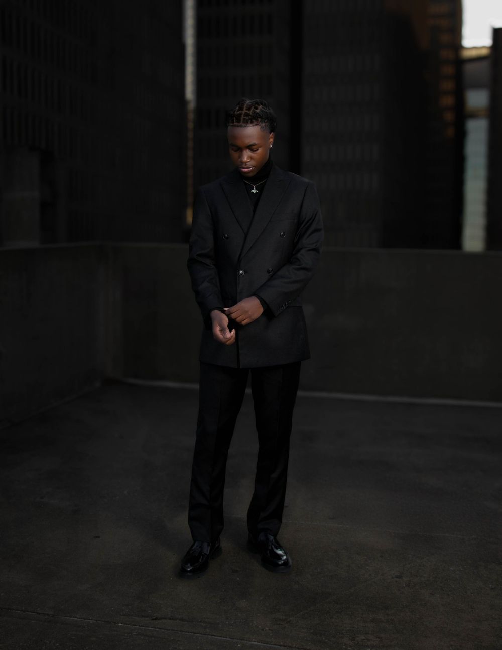 Someone wearing an all black formal outfit with blazer and dress shoes stands in a dimly lit urban environment.