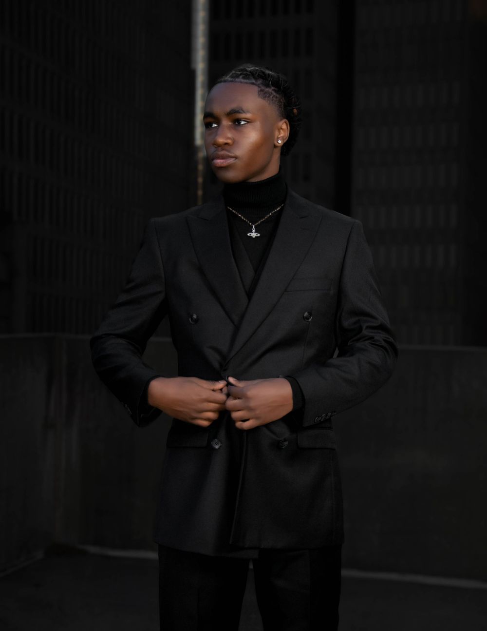 Person in all-black formal attire with a double-breasted blazer and turtle neck poses against dark background.