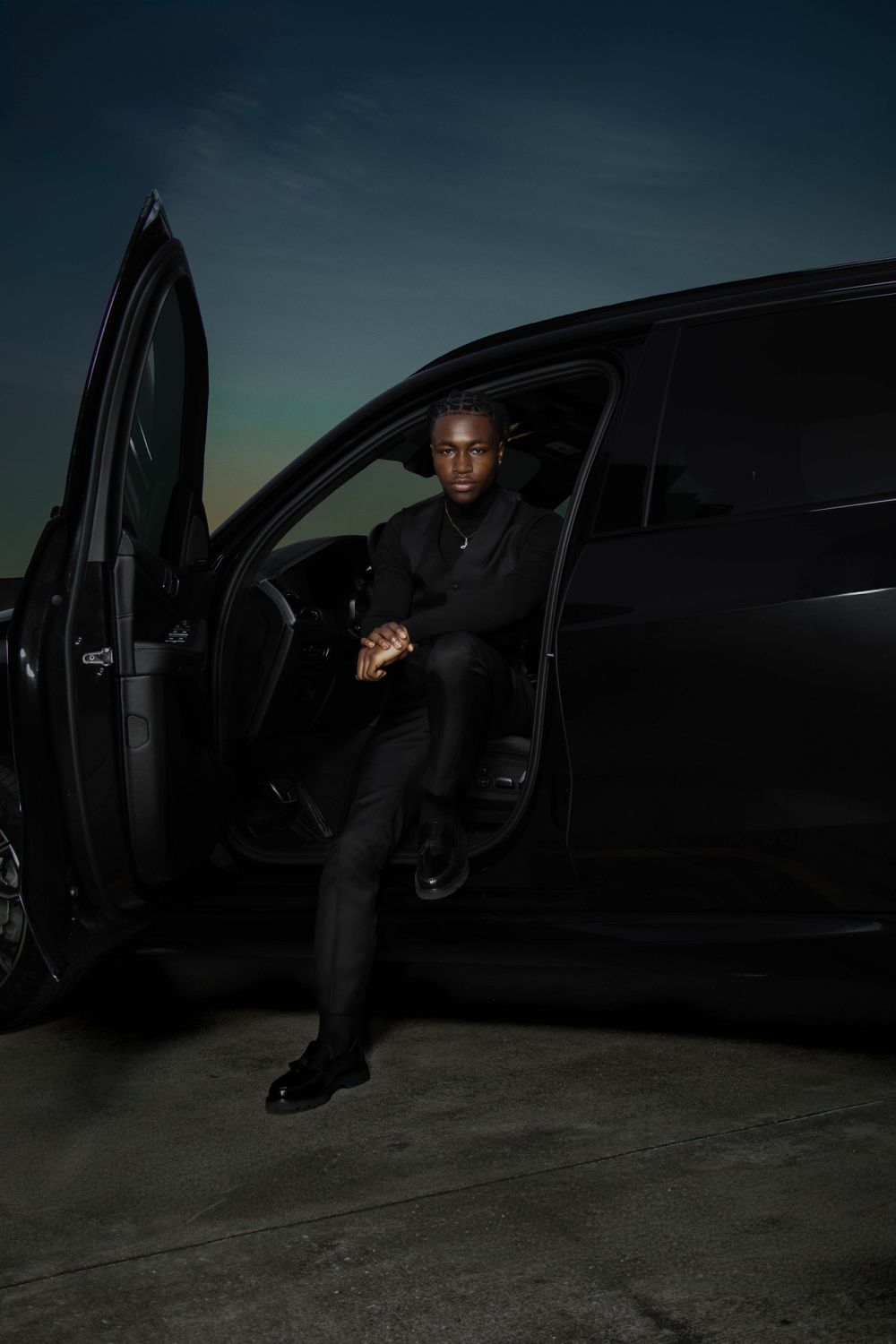 Someone in an all black outfit sits in a black luxury car at night with the door open against a dark moody sky.