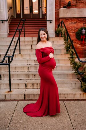 A pregnant woman in an elegant red off-shoulder gown poses on stone steps decorated with holiday greenery.