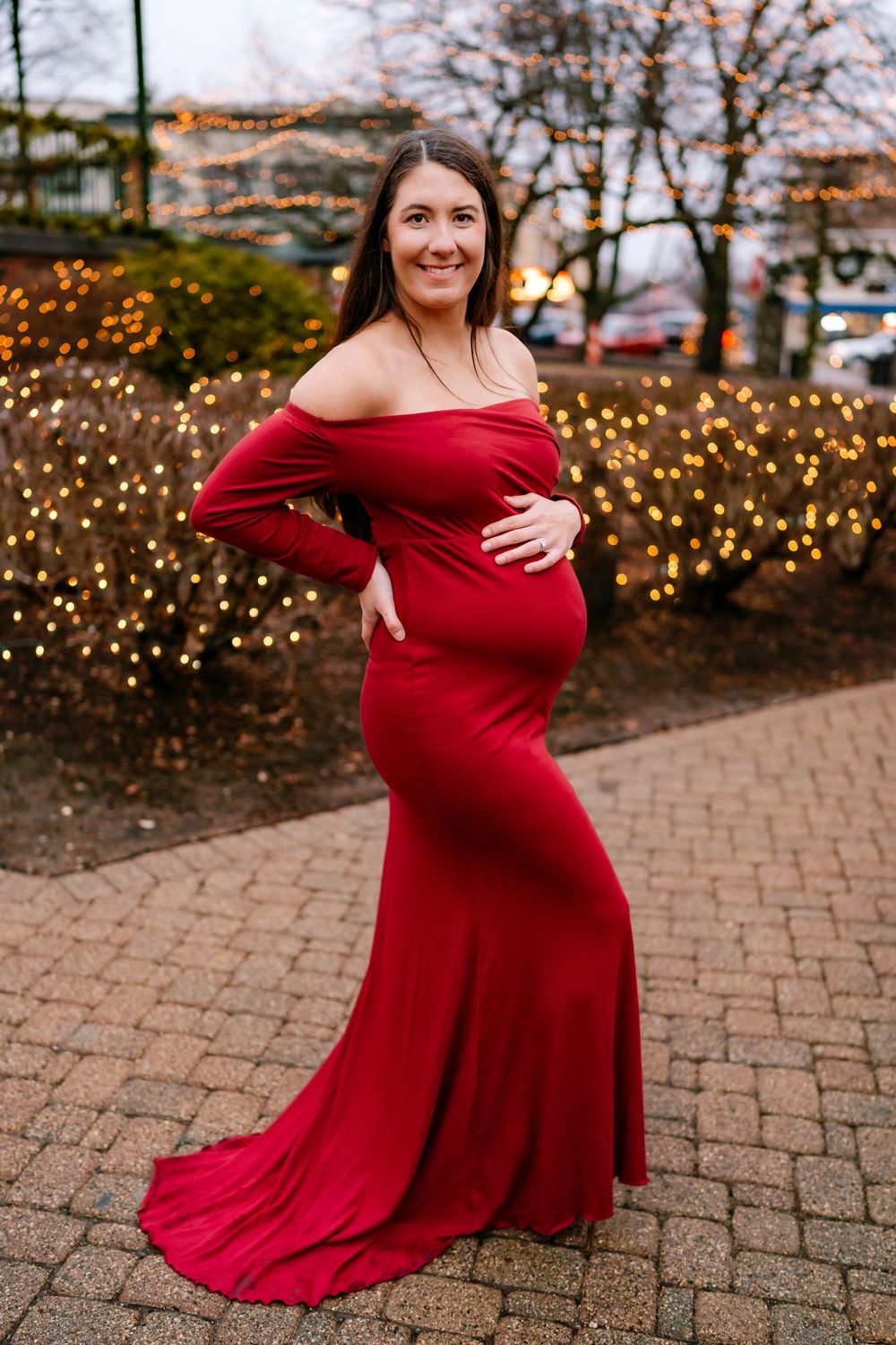 A maternity photo of an expectant mother in a flowing red off-shoulder gown standing on brick pavement with twinkle lights behind.