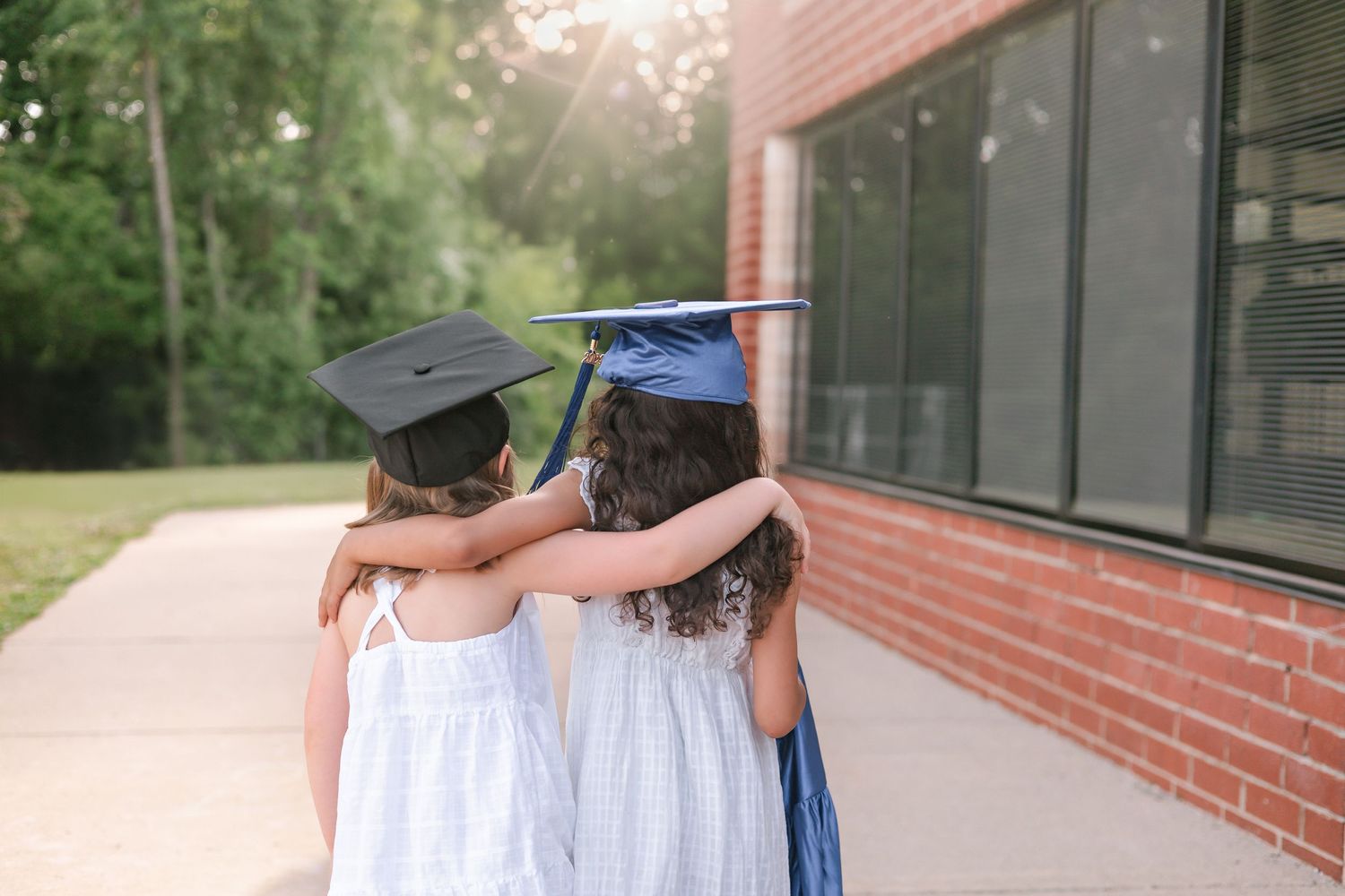 Two young girls in white dresses and graduation caps embrace each other outside a brick school building.