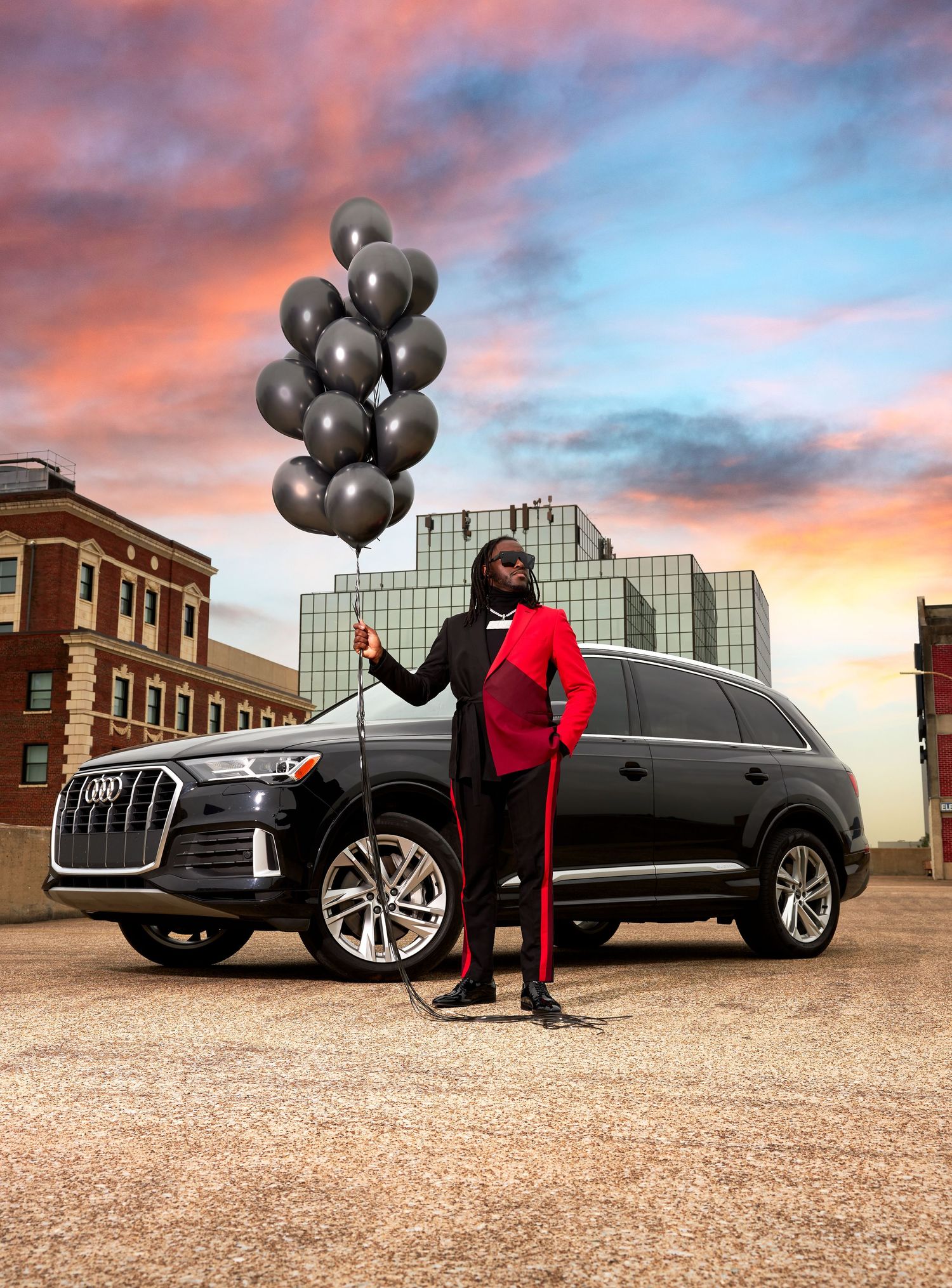 Person in red outfit holds black balloons next to luxury SUV at sunset.