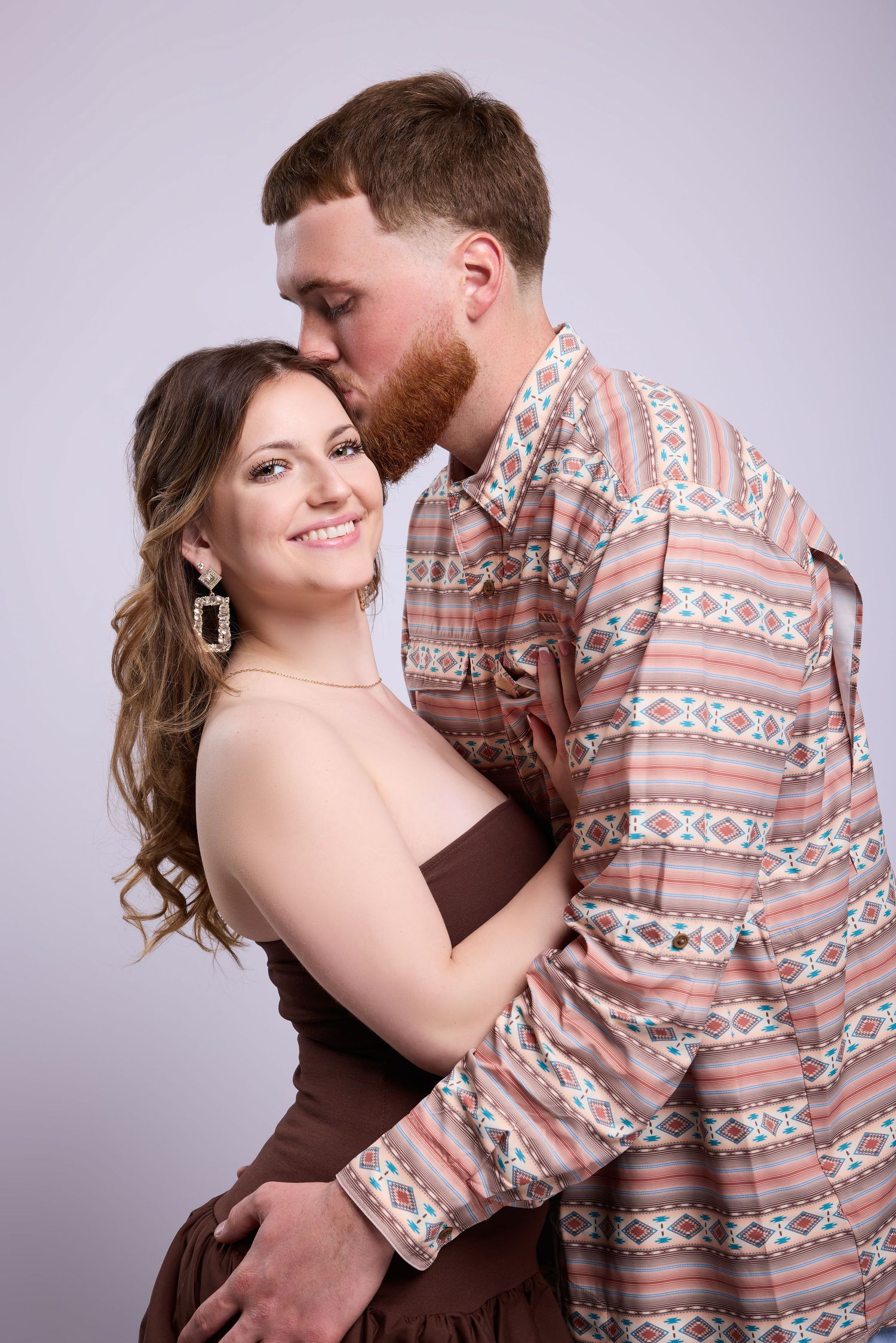 A romantic portrait of a couple embracing, with one wearing a patterned shirt and the other in a brown dress.