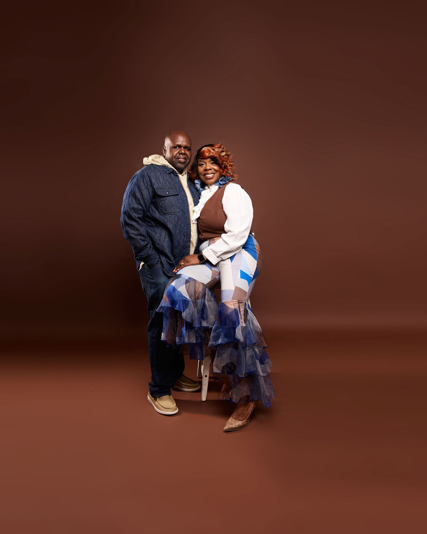 Two people pose together against a brown studio backdrop, wearing blue denim and white ruffled clothes.