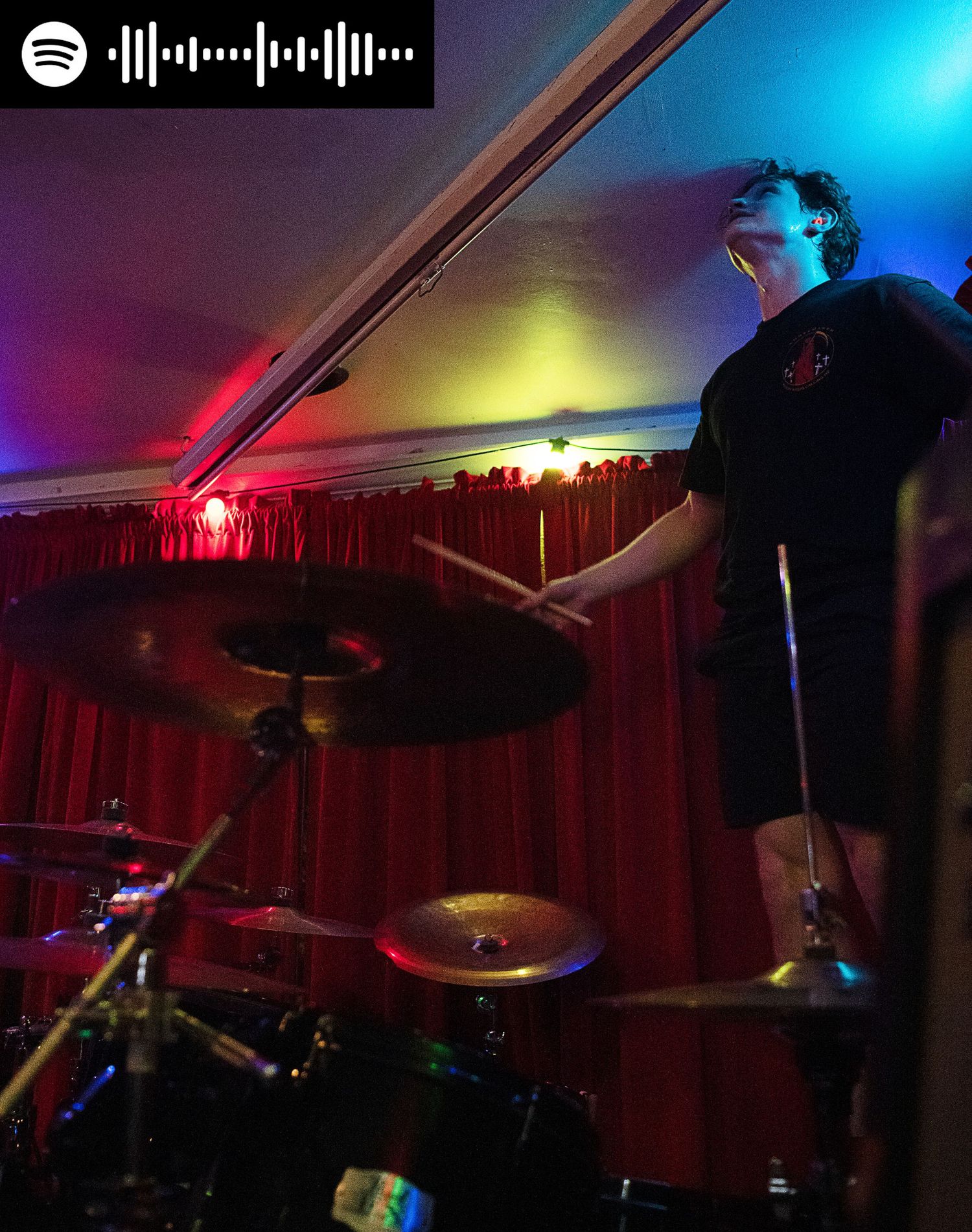 Eric of the publik standing on his drum stool holding arms out with spotify code in top left corner