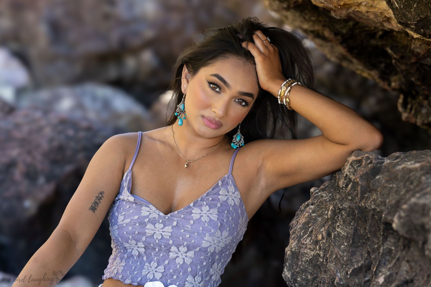 A person with dark hair wearing a purple top poses outdoors in natural sunlight.
