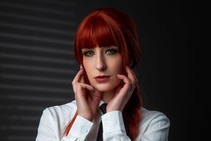 Portrait of a person with vibrant red hair and bangs wearing a white button-up blouse against a dark backdrop.