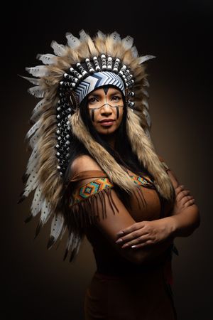A figure wearing a Native American headdress and traditional attire poses dramatically against a dark background.
